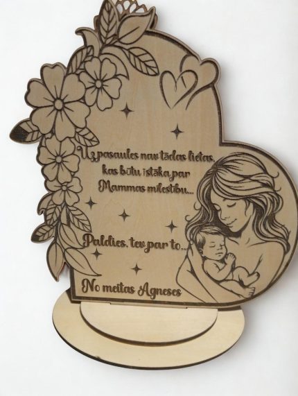 Koka dekors ar gravētu mātes un bērna ilustrāciju, ziedu ornamentiem un personalizētu tekstu “No meitas Agnese”, novietots uz stenda.