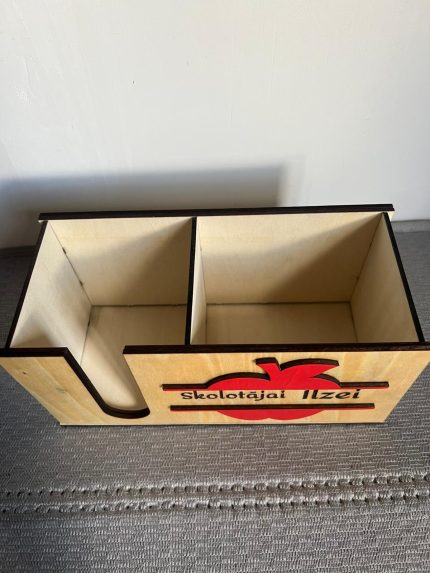 Koka galda organizators ar personalizētu uzrakstu "Skolotājai Ilzei", ar sarkanu ābola dekori un krāsainiem zīmuļiem iekšpusē, novietots uz rakstāmgalda.