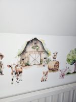 Fermas dzīvnieku līmpapīra sienas uzlīmes bērnu istabā ar govi, jēru, kazlēnu, cālīti, kaķēnu un fermas ēkām, izvietotas virs bērnu gultiņas.
