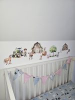 Fermas dzīvnieku līmpapīra sienas uzlīmes bērnu istabā ar govi, jēru, kazlēnu, cālīti, kaķēnu un fermas ēkām, izvietotas virs bērnu gultiņas.