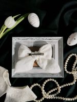 Balta lina matu bante ar rokdarba izšuvumu un cirkonija pērlītēm, metāla klipsis redzams aizmugurē, kompozīcija uz melna auduma ar pērlēm un Lieldienu olām.