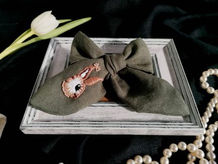 Olīvzaļa lina matu bante ar rokas izšuvumu – zaķītis ar cirkonija pērlītēm, novietota koka rāmītī uz tumša auduma fona ar pērļu virteni un tulpi.