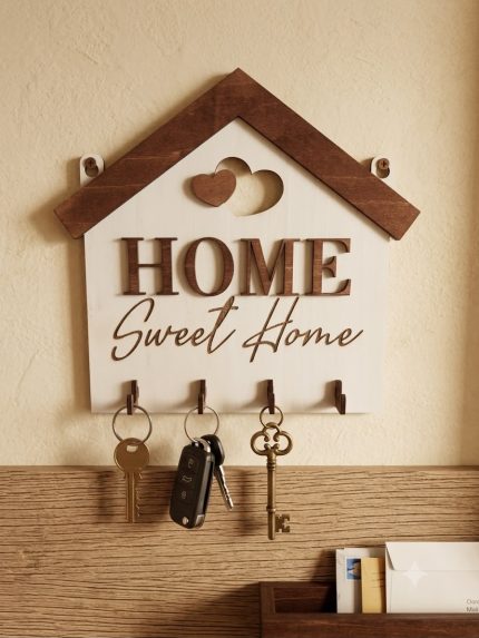 "Sienas atslēgu turētājs mājiņas formā ar uzrakstu 'Home Sweet Home' un sirsniņu dekoriem, piestiprināts pie gaišas sienas virs koka plaukta, uz kura atrodas vēstules; uz āķīšiem karājas mājas un auto atslēgas."