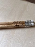 "Divi koka zīmuļi ar iegravētiem vārdiem 'Aleks' un 'Marta' novietoti uz atvērtas rūtiņu burtnīcas uz koka galda."