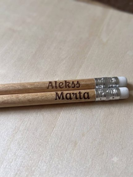 "Divi koka zīmuļi ar iegravētiem vārdiem 'Aleks' un 'Marta' novietoti uz atvērtas rūtiņu burtnīcas uz koka galda."